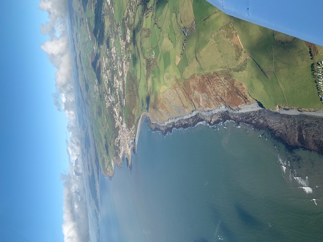 A curving coastline, green fields and a seaside town seen beyond the aircraft wing